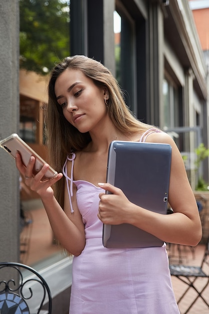 Jonge aziatische vrouw in roze jurk die buiten staat met laptop met smartphone