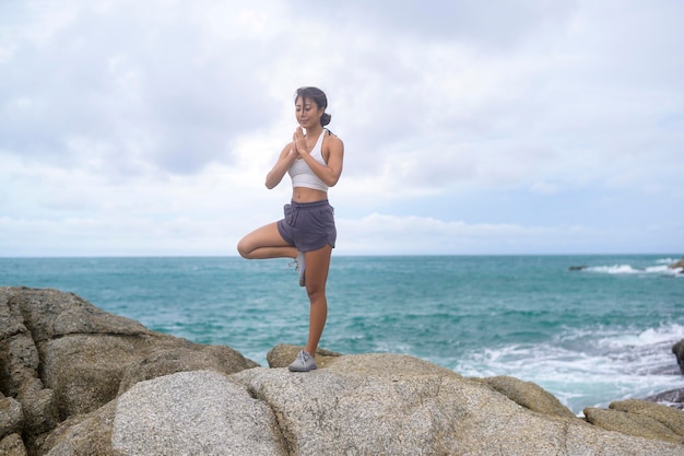 Jonge aziatische vrouw in sportkleding die yoga op de rots doet bij het concept van gezondheid en meditatie aan zee