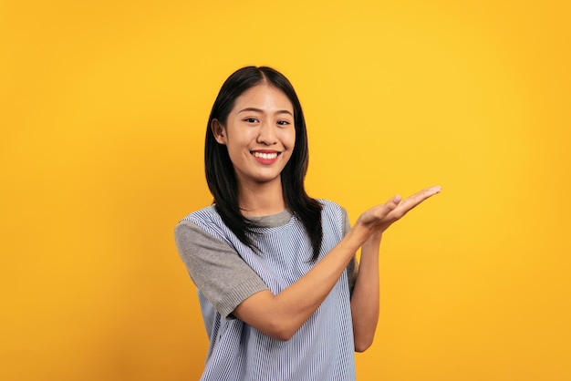 Jonge aziatische vrouw in vrijetijdskleding opent beide handen voor reclame op gele achtergrond