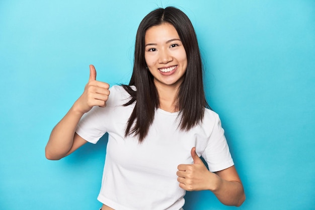 Jonge Aziatische vrouw in wit T-shirt studio shot met beide duimen omhoog glimlachend en zelfverzekerd