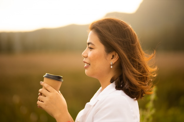 Jonge Aziatische vrouw in witte overhemd het drinken koffie openlucht in de tijd van de zonsopgangzomer