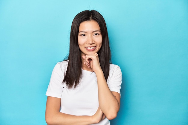 Jonge Aziatische vrouw in witte t-shirtstudio-opname glimlachend, blij en zelfverzekerd, kin met de hand aanrakend