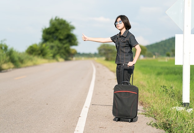 Jonge Aziatische vrouw kort haar en het dragen van een zonnebril met bagage liften langs een weg en duimen omhoog in landweg Thailand