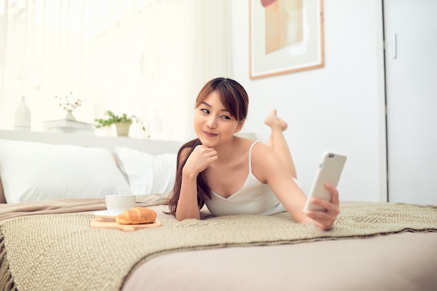 Jonge Aziatische vrouw liggend op bed en met behulp van mobiele telefoon selfie in haar huis.