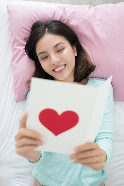 Jonge aziatische vrouw liggend op bed, met wenskaart met rode hartvorm.