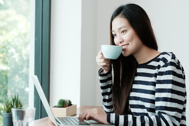 Jonge Aziatische vrouw met behulp van laptopcomputer en koffie drinken op café achtergrond