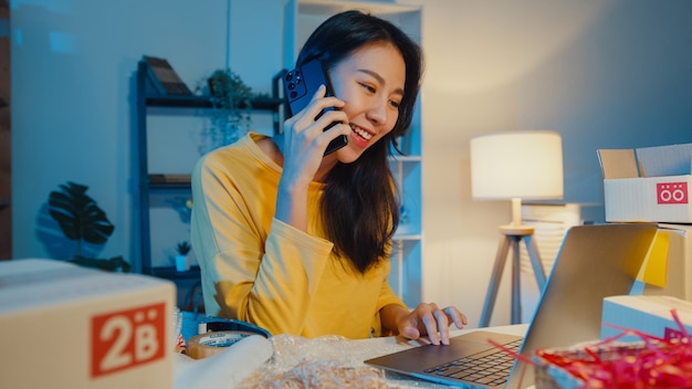 Jonge Aziatische vrouw met behulp van smartphone praten met klant voor cheque bevestigen bestelling in voorraad op laptop