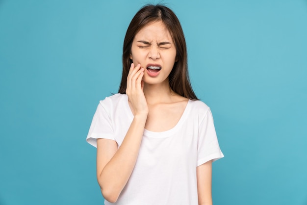 Jonge Aziatische vrouw met beugels met gevoelige tanden en handen raakt de wangen aan op een blauwe achtergrond, Concept mondhygiëne en gezondheidszorg.