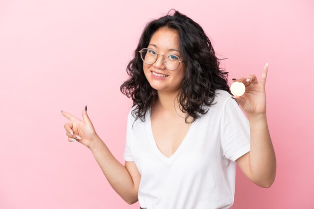 Jonge aziatische vrouw met een Bitcoin geïsoleerd op een roze achtergrond die met de vinger naar de zijkant wijst