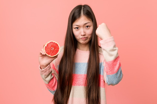 Jonge Aziatische vrouw met een grapefruit die in agressieve gelaatsuitdrukking toont.