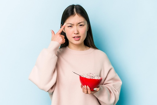 Jonge aziatische vrouw met een kom granen geïsoleerd op een blauwe achtergrond met een teleurstellingsgebaar met wijsvinger