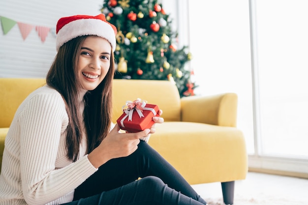 Jonge Aziatische vrouw met een trui en een kerstmuts glimlacht en houdt een kerstcadeau vast in een rode doos met lint zittend in een woonkamer met een versierde kerstboom op de achtergrond