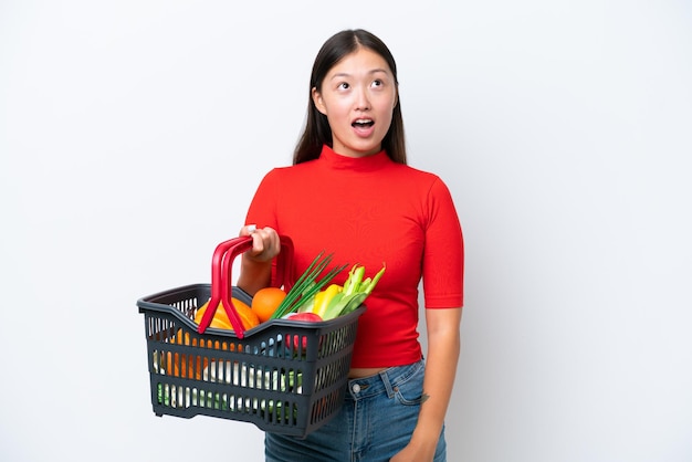 Jonge Aziatische vrouw met een winkelmandje vol voedsel geïsoleerd op een witte achtergrond, omhoog kijkend en met een verbaasde uitdrukking
