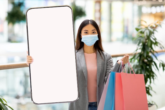 Jonge aziatische vrouw met gezichtsmasker met cadeauzakjes en mobiele telefoon met mockup bij supermarkt online