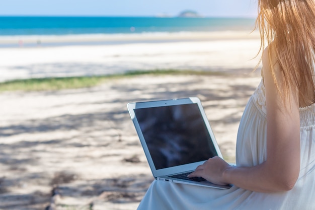 Jonge Aziatische vrouw met laptop in jurk zittend op het strand, meisje Freelancer werken