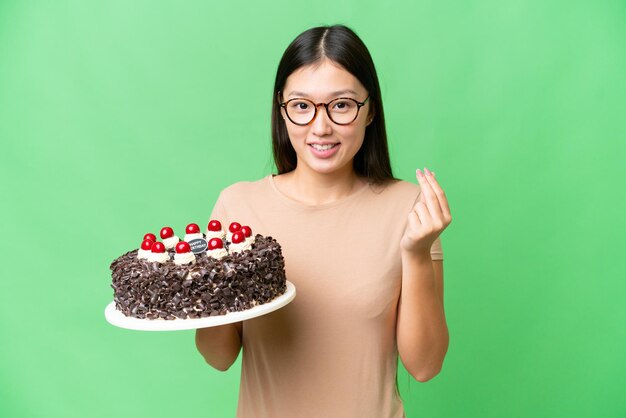 Jonge Aziatische vrouw met verjaardagstaart over geïsoleerde chroma key achtergrond die geldgebaar maakt