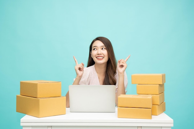Jonge Aziatische vrouw opstarten kleine bedrijven freelance wijzende vinger omhoog met pakketdoos en computer laptop op tafel en zittend geïsoleerd op groene achtergrond, Online marketing en levering concept