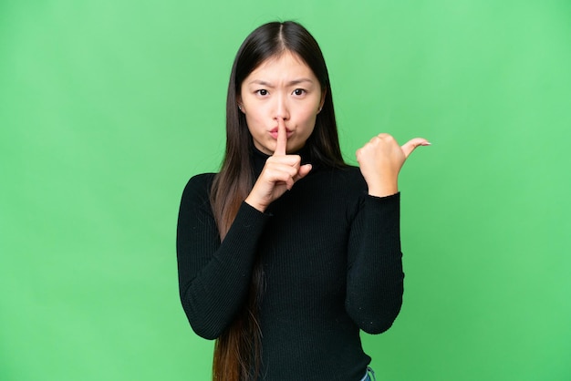 Jonge Aziatische vrouw over geïsoleerde chroma key-achtergrond die naar de zijkant wijst en stiltegebaar doet