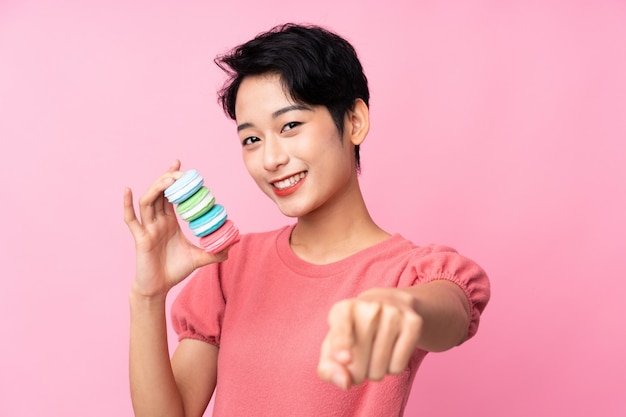 Jonge aziatische vrouw over geïsoleerde roze muur die kleurrijke franse macarons en puntenvinger op u houdt