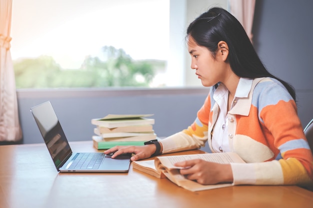 Jonge Aziatische vrouw student in studentenuniform studeren, lezen van een boek, laptop in universiteits- of hogeschoolbibliotheek. Jeugdmeisjesstudent en bijlesonderwijs met een technologisch leerconcept