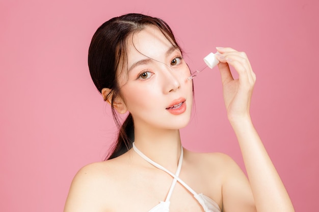 Jonge Aziatische vrouw verzameld in paardenstaart met natuurlijke make-up heeft dikke lippen en een schone, frisse huid, gekleed in een wit hemdje dat hydraterend serum op het gezicht aanbrengt op een geïsoleerde roze achtergrond
