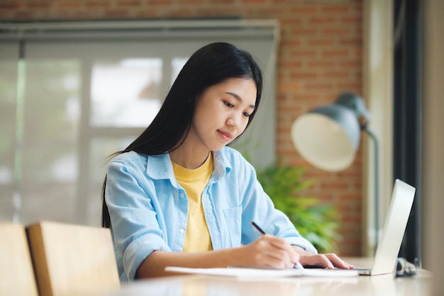 Jonge aziatische vrouw zittend aan tafel studeren en schrijven op notebook