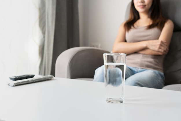 Jonge Aziatische vrouw zittend op de bank terwijl ze tv kijkt in de woonkamer thuis met een glas water op bureau Ontspan thuis gezonde levensstijl concept Kopieer ruimte