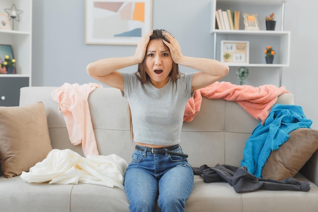 Jonge aziatische vrouw zittend op een bank kijkend naar puinhoop in haar woonkamer bij verspreide was in paniek, hand in hand op haar hoofd, bezorgd en gestrest