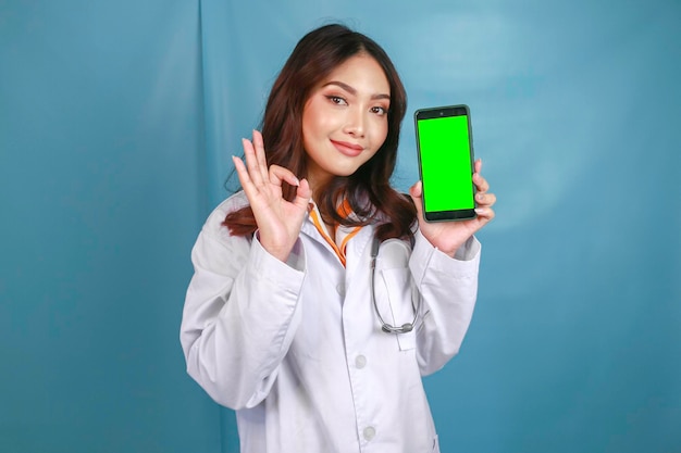 Jonge Aziatische vrouwelijke arts wijst en toont groen scherm of kopieert ruimte op haar smartphone met duimen omhoog of OK-teken