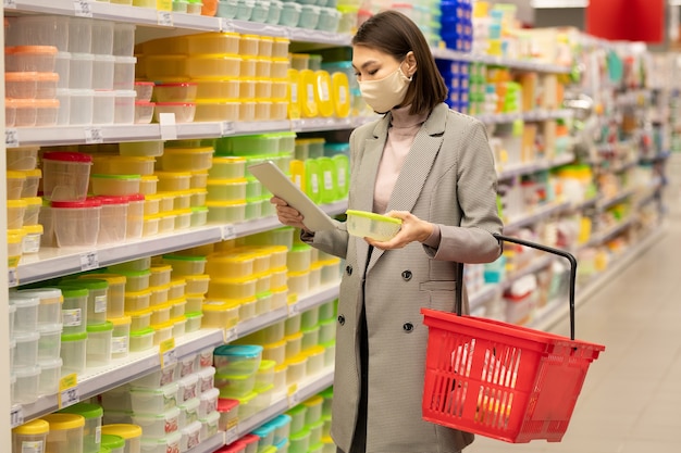 Jonge Aziatische vrouwelijke consument met rode winkelmand die bij een grote plank met containers staat en door hun kenmerken in tablet kijkt