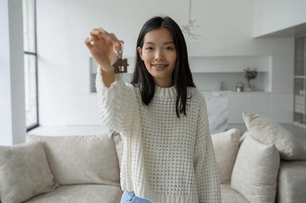 Jonge Aziatische vrouwelijke koper-huurder kijkt in de camera met sleutels in haar hand een gelukkige vrouw trots op