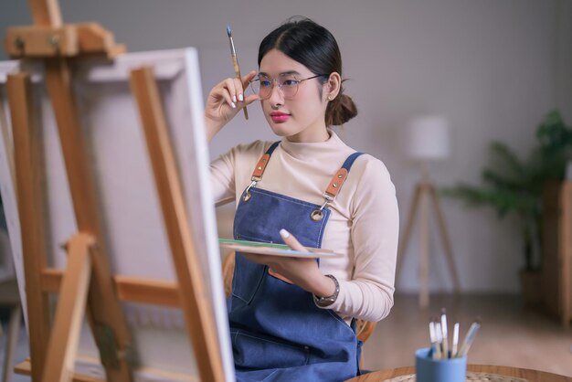 Foto jonge aziatische vrouwelijke kunstenaar die een bril aanraakt en naar het doek kijkt om te denken idee schilderen kunstwerk