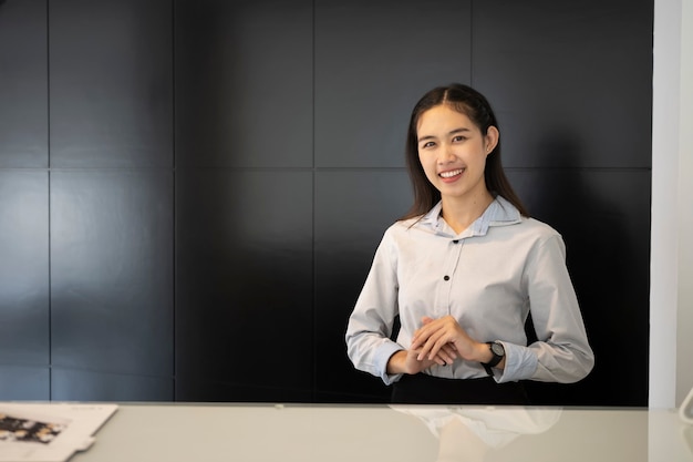 Jonge aziatische vrouwelijke receptioniste die bij de receptie staat en glimlacht om bezoekers in het hotel te verwelkomen.