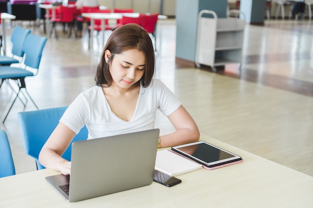 Jonge aziatische vrouwelijke student die met laptop, tablet, telefoon en notitieboekje bij bibliotheek werkt.