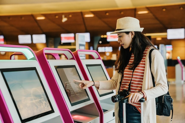 jonge Aziatische vrouwelijke zakenreiziger met behulp van selfservice check-in machine in de lobby van de luchthaven binnen. meisjesfotograaf met cameravinger die het scherm aanraakt en kiest voor het kiezen van een automatisch tellerconcept.