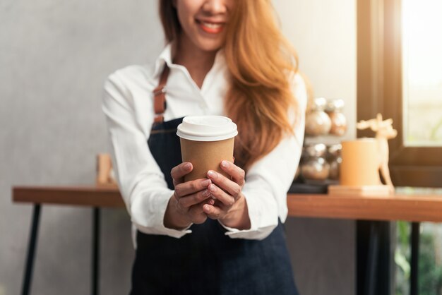 Jonge Aziatische vrouwenbarista die een beschikbare koffiekop met het glimlachen gezicht houden bij koffieachtergrond