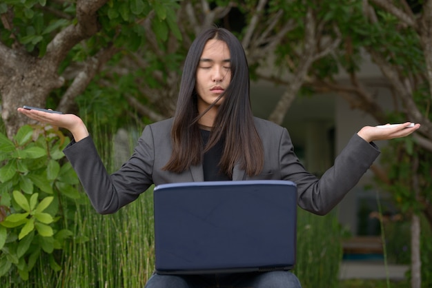 Jonge Aziatische zakenman die met lang haar twee telefoons en laptop houdt tijdens het mediteren in het park