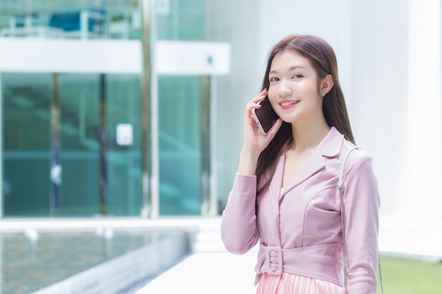 Jonge Aziatische zakenprofessional in roze jurk staat buiten en kijkt naar de camera belt de telefoon