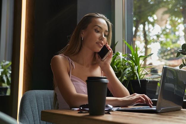 Jonge aziatische zakenvrouw die notities schrijft in een notitieboekje in café dat op een laptop werkt