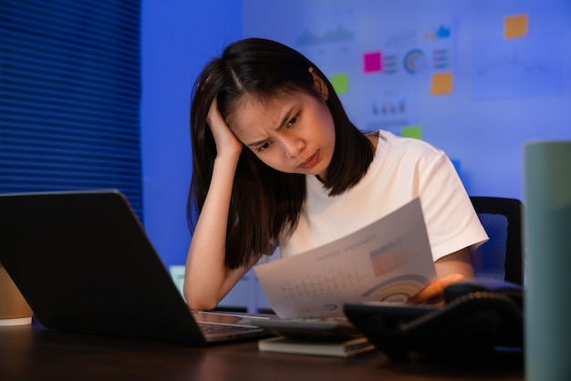 Jonge Aziatische zakenvrouw die op laptop werkt met hoofdpijn omdat ze 's nachts op kantoor migraine heeft.