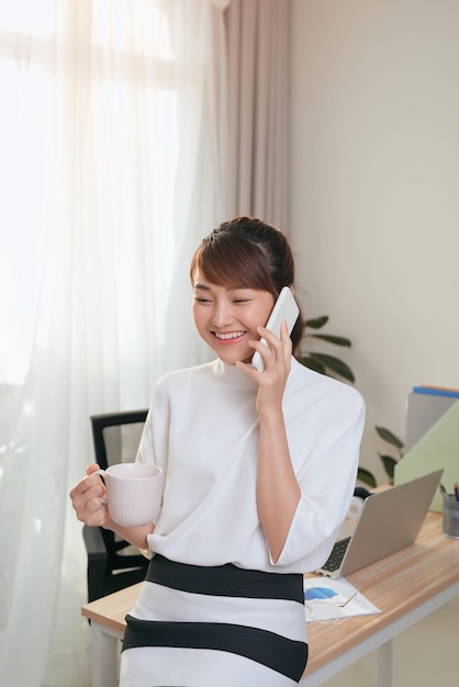 Jonge Aziatische zakenvrouw die telefonisch praat terwijl ze koffie drinkt aan tafel in kantoor