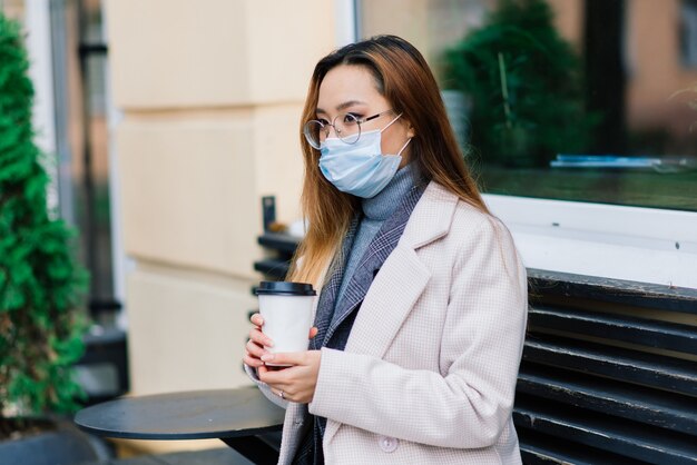 Jonge Aziatische zakenvrouw met masker die telefoon gebruikt en met afstand buiten de coffeeshop zit