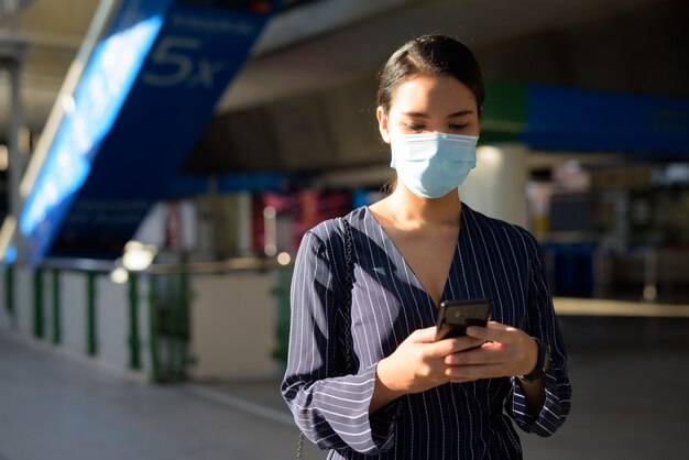 Jonge Aziatische zakenvrouw met masker met behulp van telefoon tijdens het weglopen van het treinstation