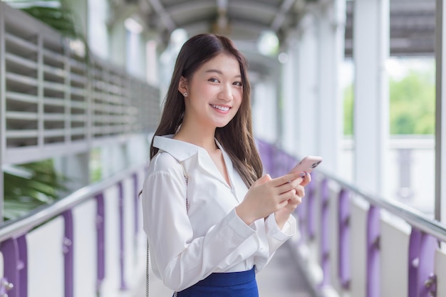 Jonge Aziatische zakenvrouw staat op een viaduct van skytrain in de stad terwijl ze haar smartphone gebruikt