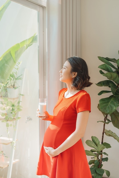 Jonge Aziatische zwangere vrouw met melkglas wanneer ze achter het raam staat.