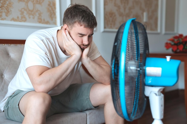 Jonge baarde man die thuis een elektrische ventilator gebruikt en op de bank zit om zich af te koelen tijdens het hete weer