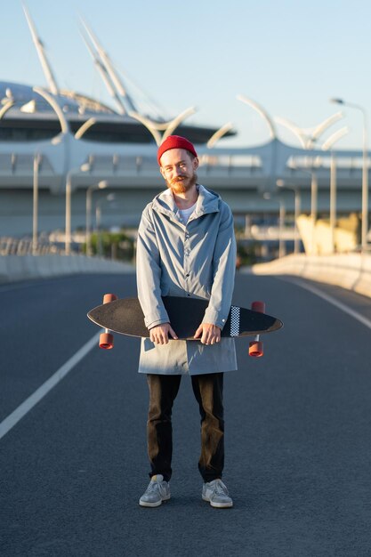 Foto jonge baardige hipster man in rode hoed houdt longboard in stadsstraat trendy stedelijke levensstijl concept