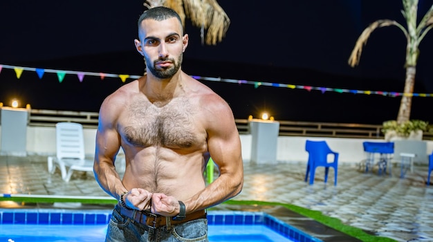 Foto jonge baardige man die tijdens zijn vakantie zijn spieren voor het zwembad laat zien met een zelfverzekerd gezicht en poseert