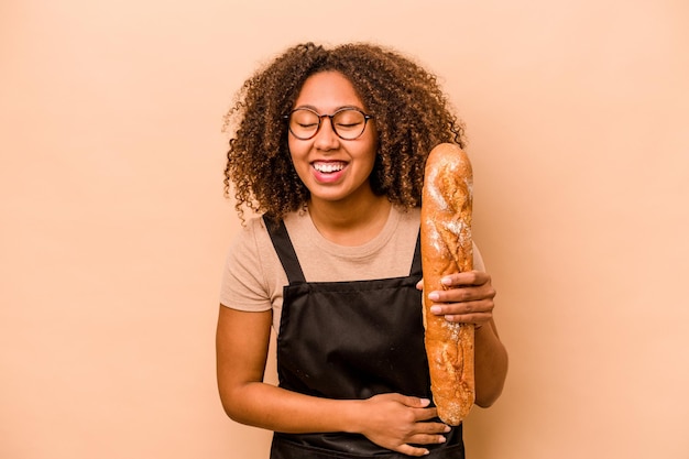Jonge bakker Afrikaanse vrouw met een brood geïsoleerd op beige achtergrond lachen en plezier hebben