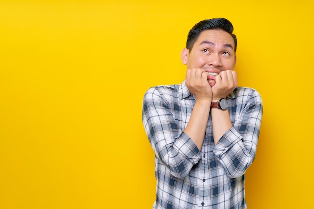 Jonge bange Aziatische man in geruite hemd die camera kijkt en zijn nagels bijt en vingers geïsoleerd op gele achtergrond mensen levensstijl concept